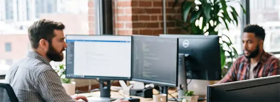 two men at desks near a window in an open office space.  They have two large monitors on their desk.